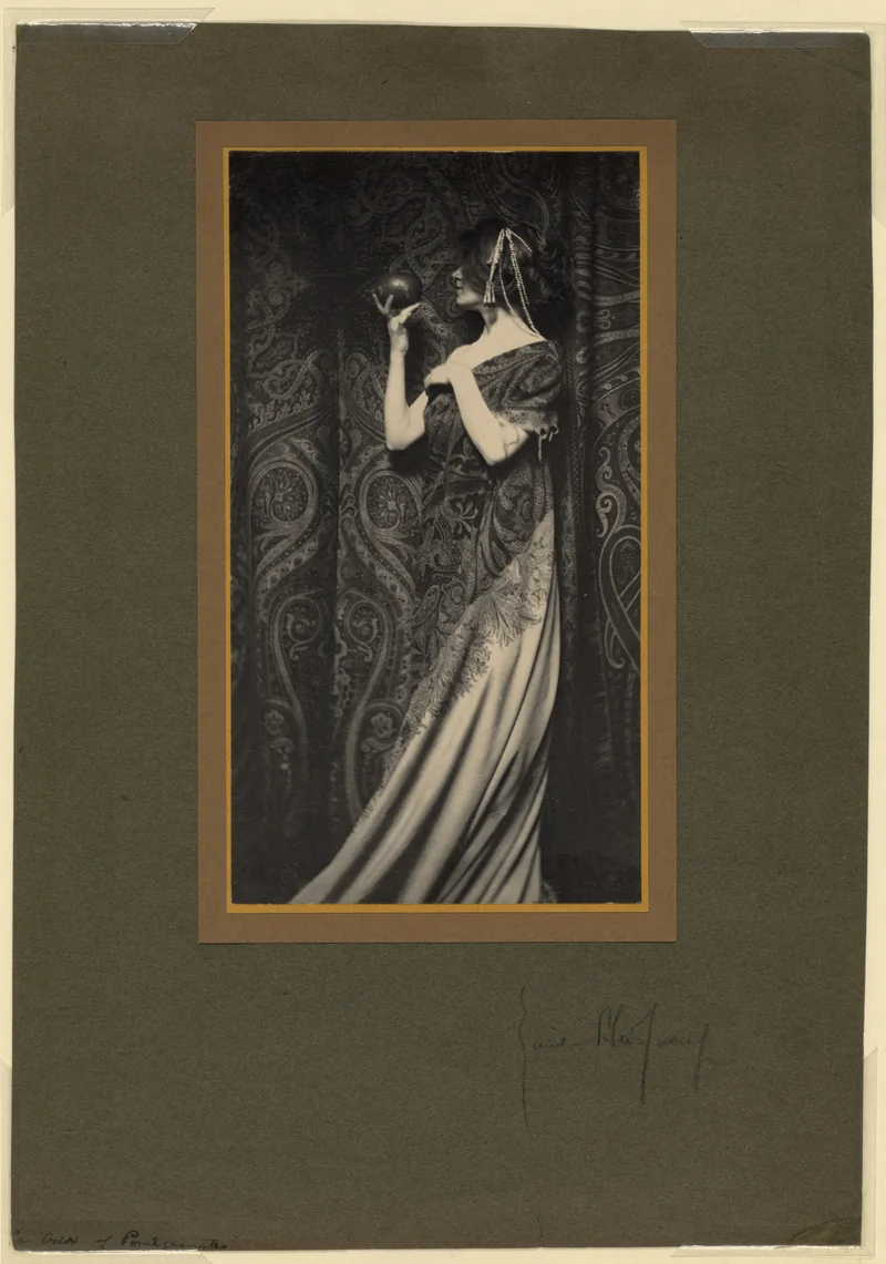 Monochrome photograph of a young woman in a light robe leaning towards a branch of pomegranates, her eyes lowered.