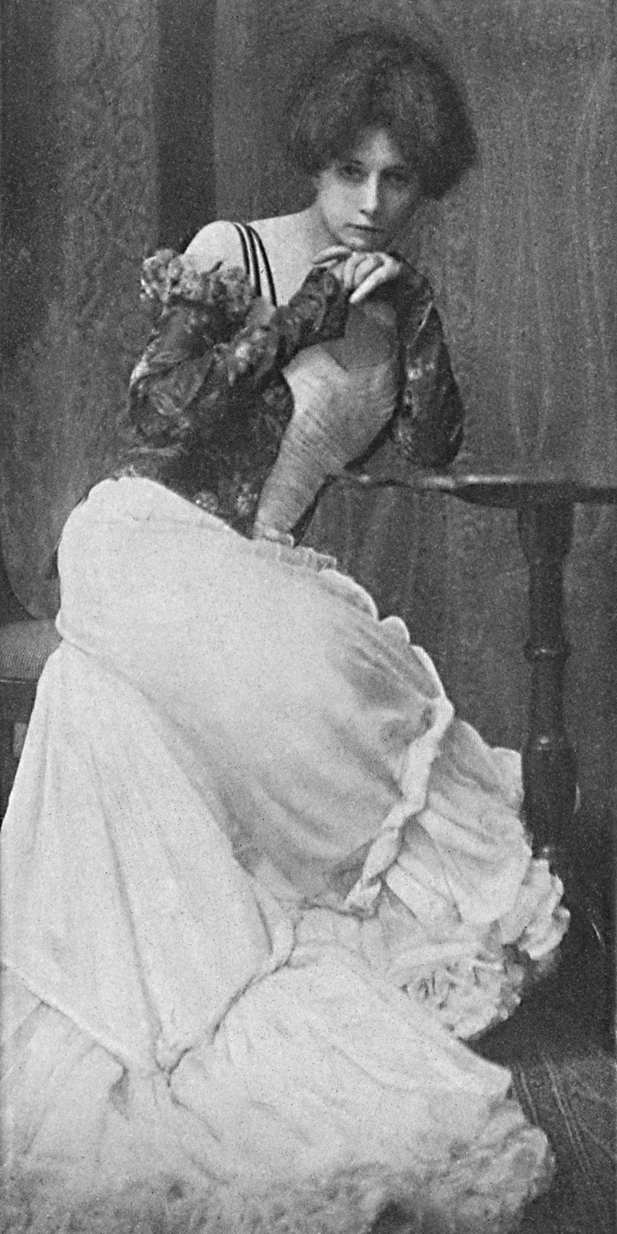 Vertical monochrome self-portrait of Zaida Ben-Yusuf, standing in profile in a long dark dress, her face turned towards the camera against a plain studio background.